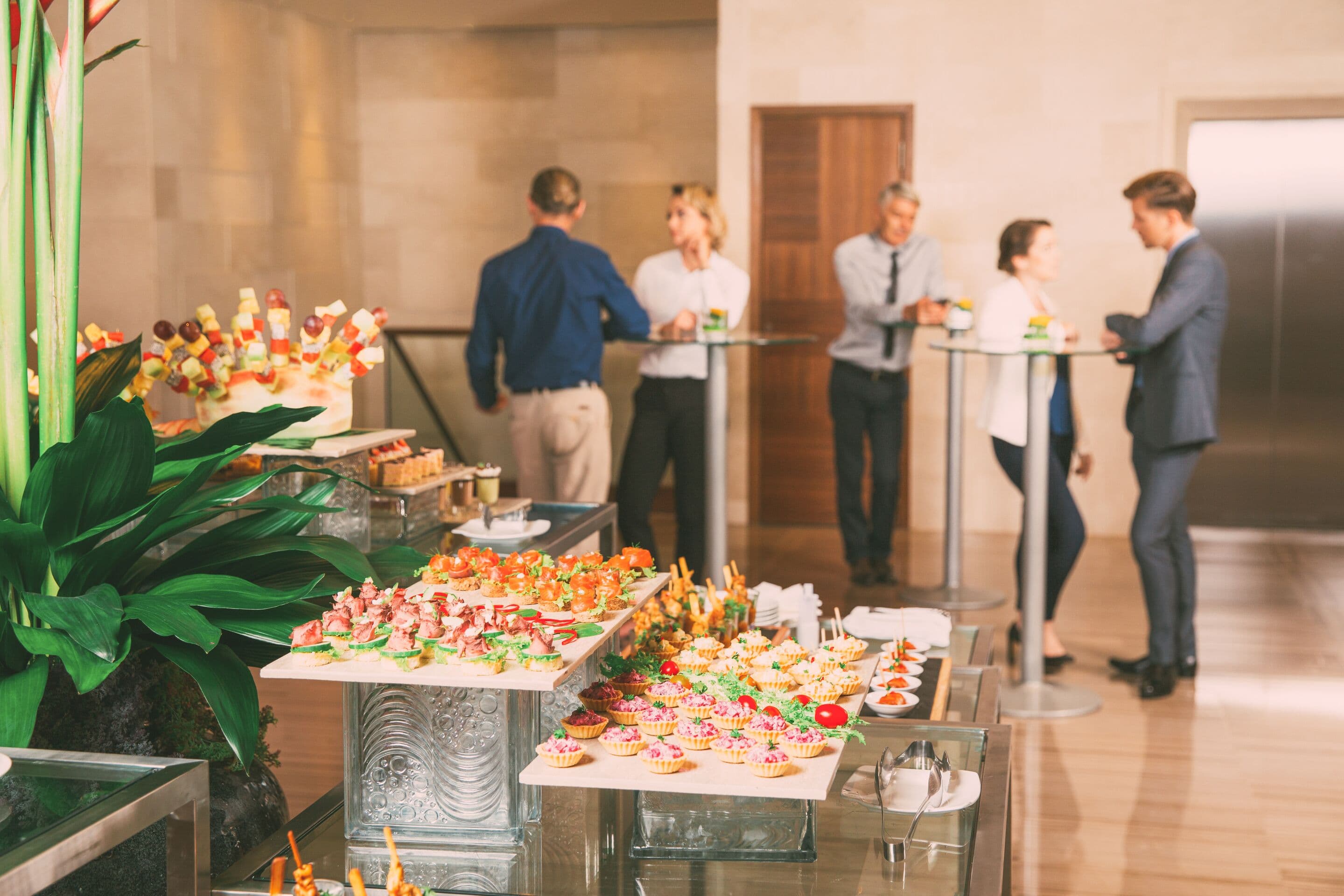 View of different canapes, tartlets and snacks on buffet table in restaurant. Business people eating and talking in background. Catering concept
